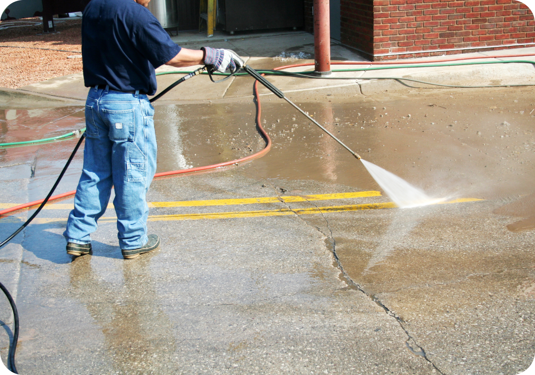 Worker using a pressure washer to clean a concrete surface with yellow road markings, near a brick building. The Benefits of Getting Your Business Pressure Cleaned