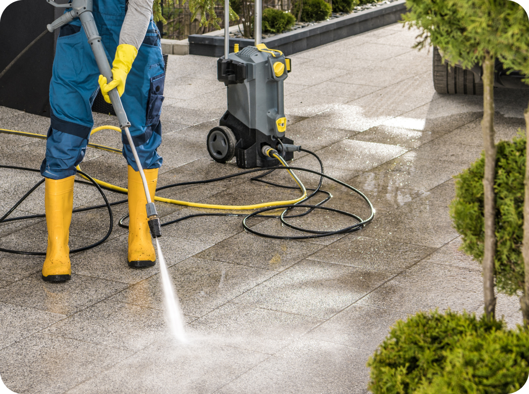 Worker in yellow boots and gloves using a pressure washer to clean a tiled outdoor surface with a pressure washer machine nearby, The Benefits of Getting Your Home Pressure Cleaned
