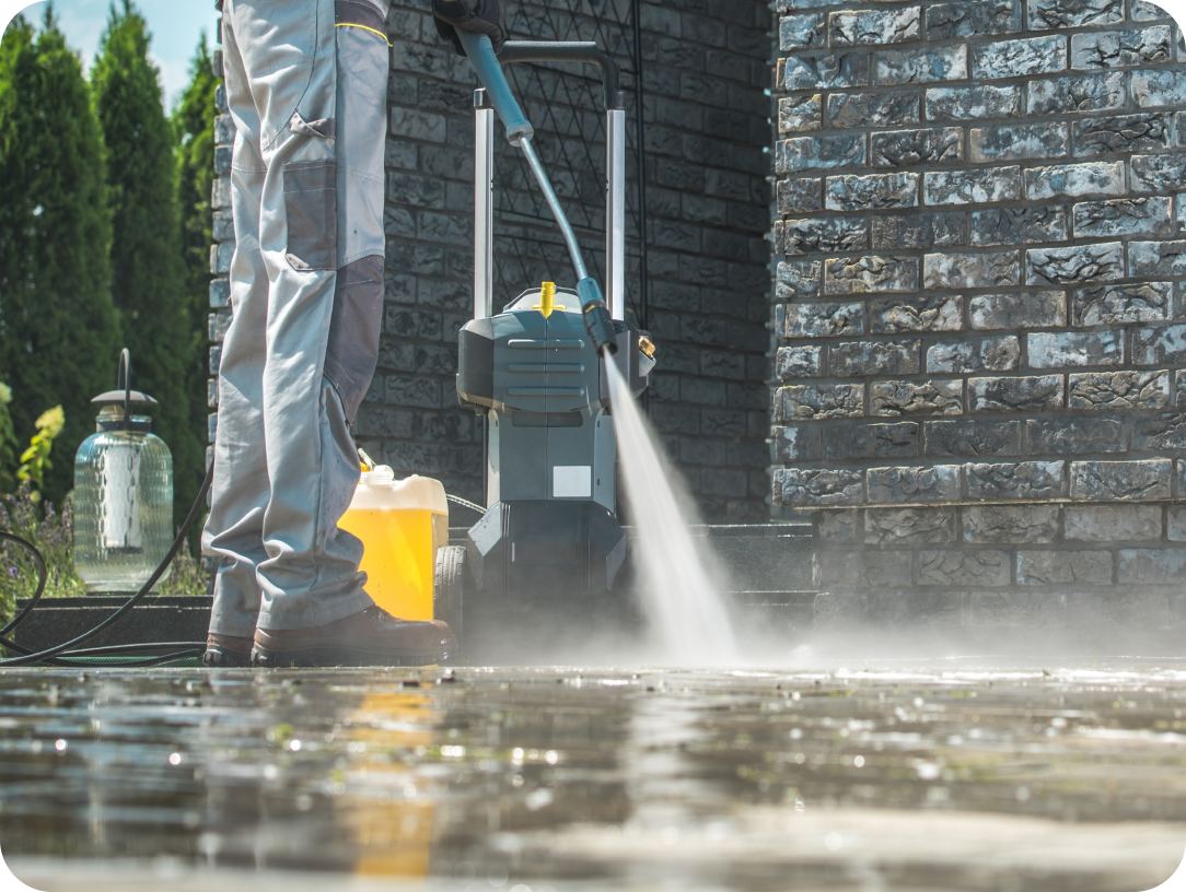 Worker using a pressure washer to clean a wet patio near a brick wall, with a pressure washer machine and cleaning supplies in the background.