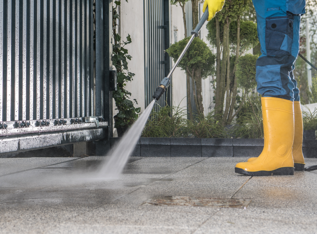 Worker in yellow boots using a pressure washer to clean a paved driveway near a metal gate, with plants and a building in the background.
