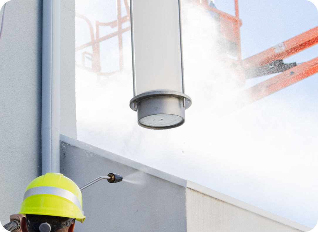 Industrial worker cleaning a vertical surface with a pressure washer, targeting a cylindrical vent or fixture, with an aerial lift visible in the background.