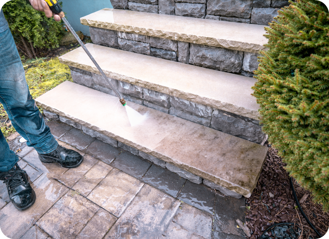 A person pressure washing a set of stone stairs, removing dirt and grime, with water spraying from a nozzle, surrounded by a landscaped outdoor area.
