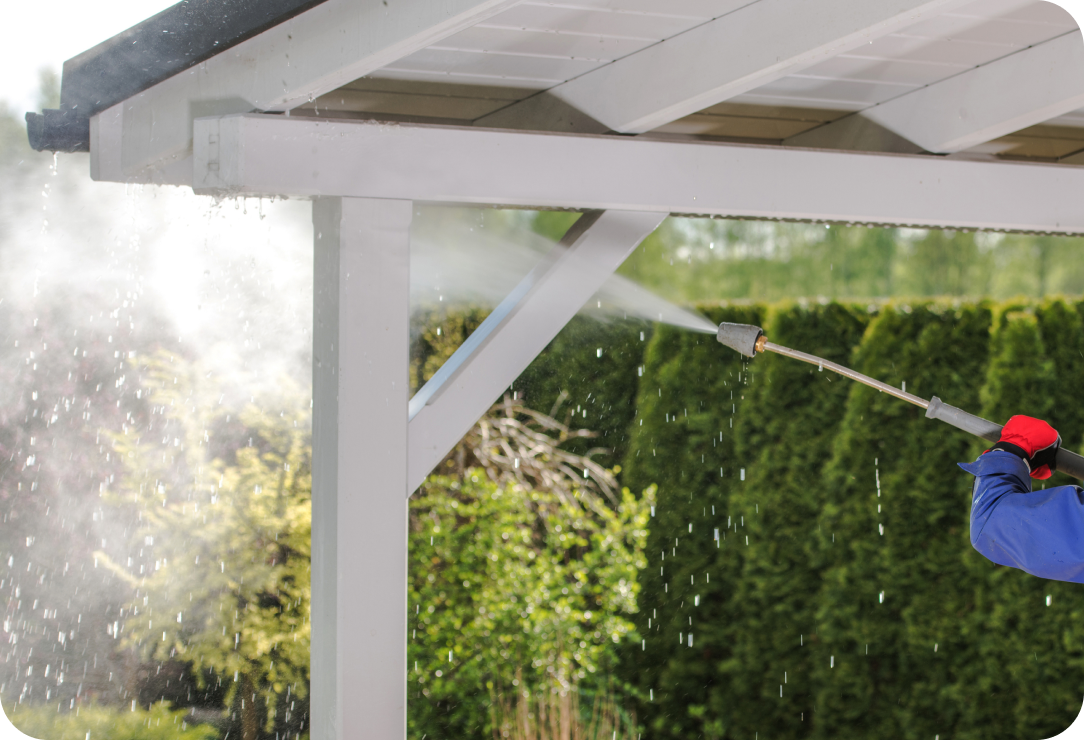 Worker using a pressure washer to clean the underside of a white pergola or patio roof, with greenery in the background.