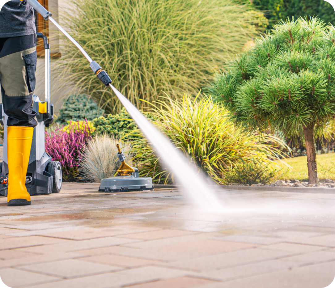 Person in yellow boots using a pressure washer to clean a residential patio surrounded by green and colorful landscaping.