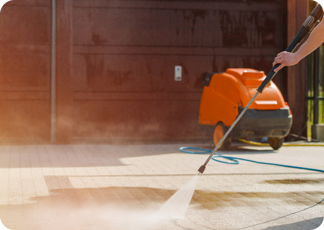 A person using a pressure washer to clean a tiled outdoor surface, with an orange industrial cleaning machine and a hose visible in the background.