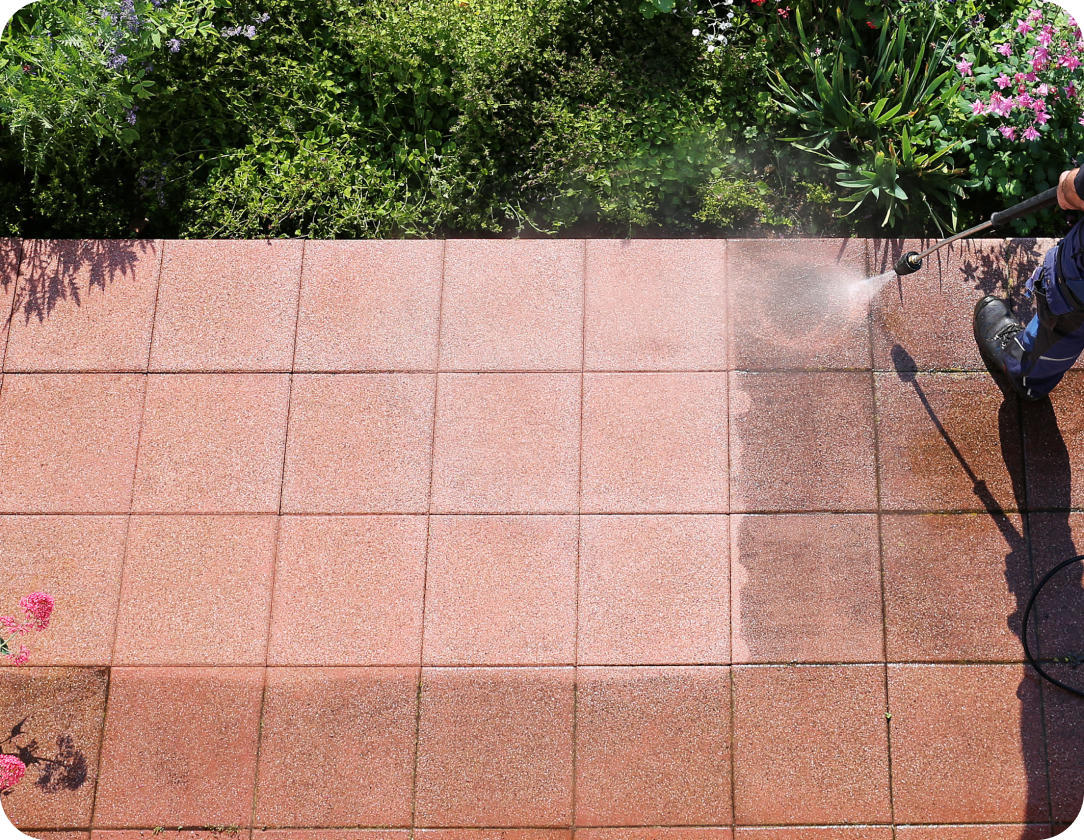 Worker using a pressure washer to clean red tiled pavement, with vibrant greenery and flowers in the background, Pressure Cleaning Services for Homes of All Sizes