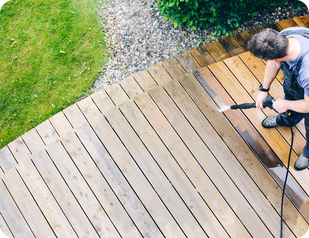 Man using a pressure washer to clean a wooden deck, with grass and gravel landscaping in the background, Signs That Your Home Needs to Be Pressure Cleaned