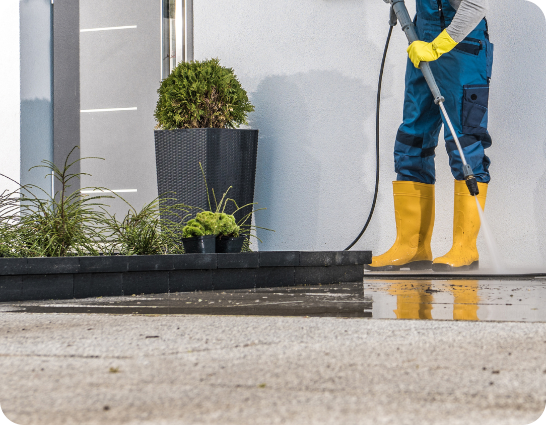 Worker in yellow boots using a pressure washer to clean the entrance area of a modern house, with potted plants and a wet pavement in the foreground.