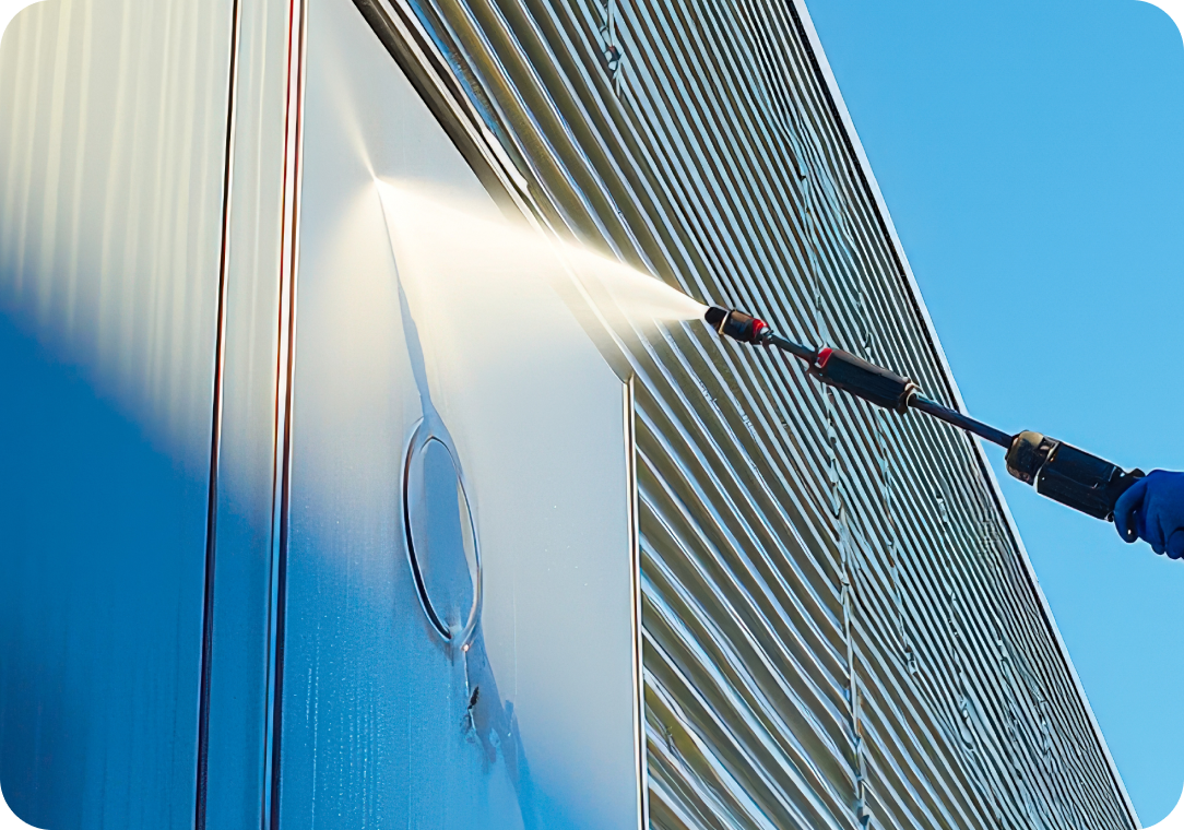 Person pressure washing the side of a modern building, with water spray visible against a reflective metal surface and a clear blue sky in the background.