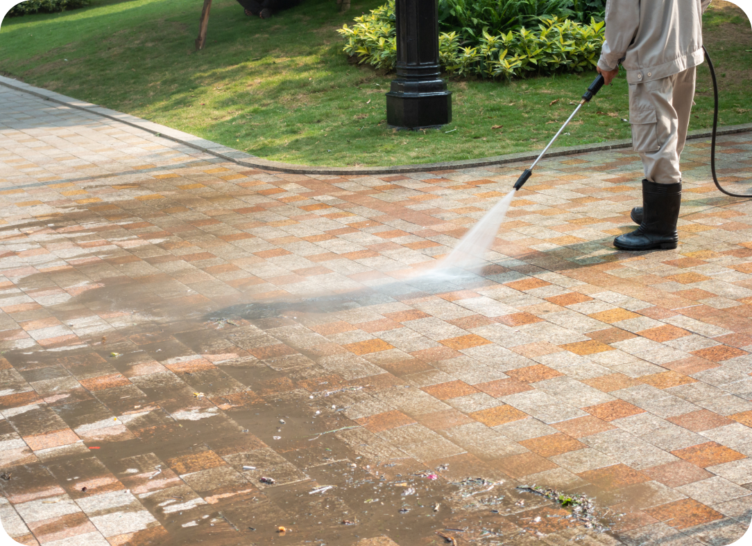 Worker using a pressure washer to clean a paved walkway with grass and shrubs in the background.