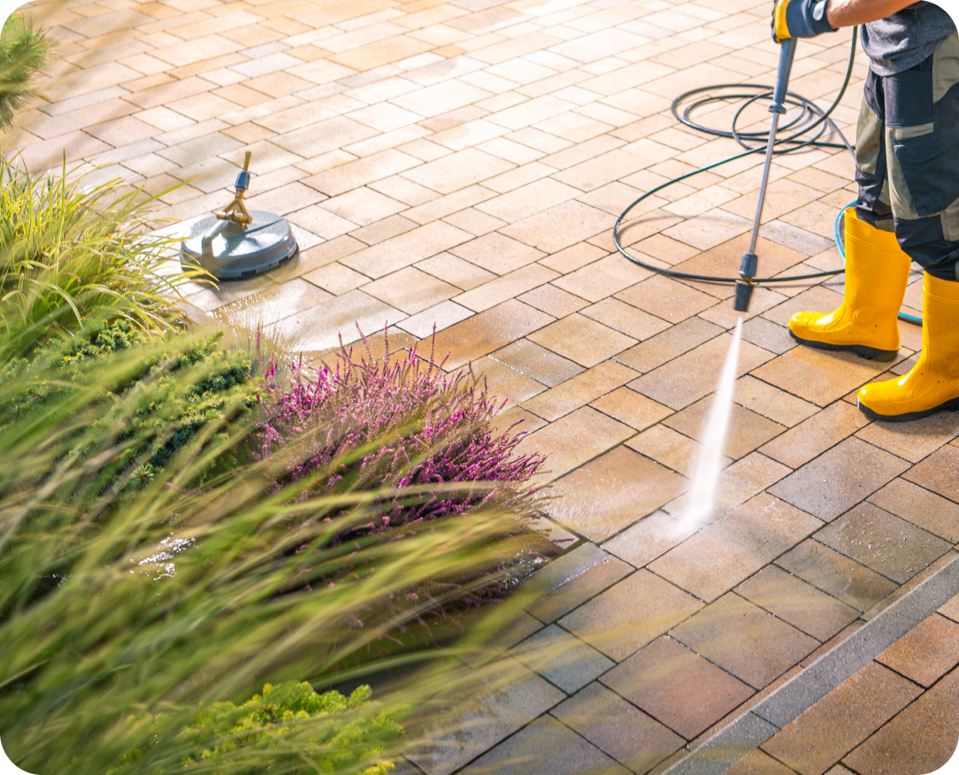 Worker wearing yellow boots using a pressure washer to clean a stone patio near vibrant landscaping with purple and green plants.